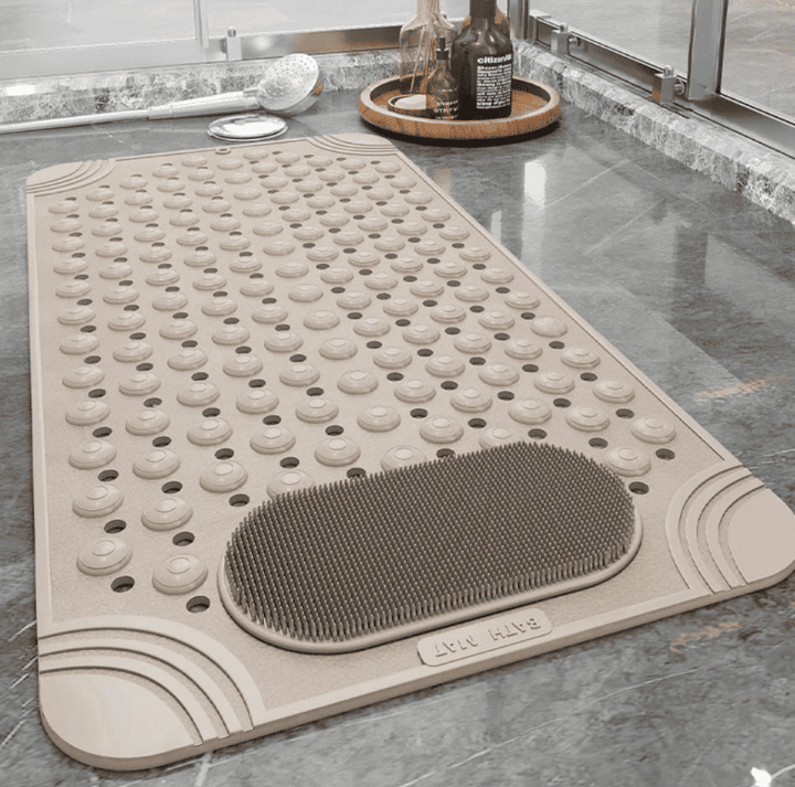 Tapis de bain antidérapant beige à picots massants et perforations d’évacuation, posé sur un carrelage gris près d’une douche.