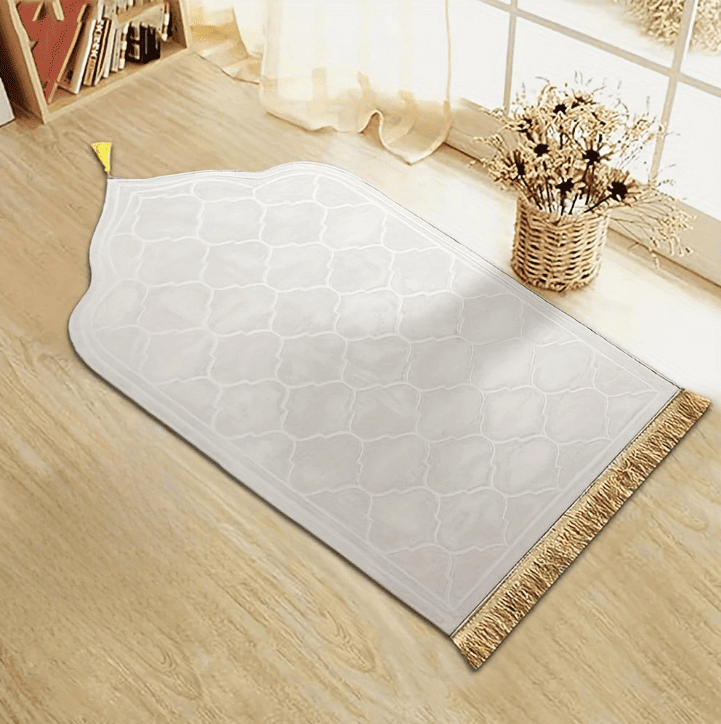 Tapis de prière épais de couleur blanche aux motifs géométriques en relief et franges dorées, déployé sur un sol en bois clair près d’une fenêtre.