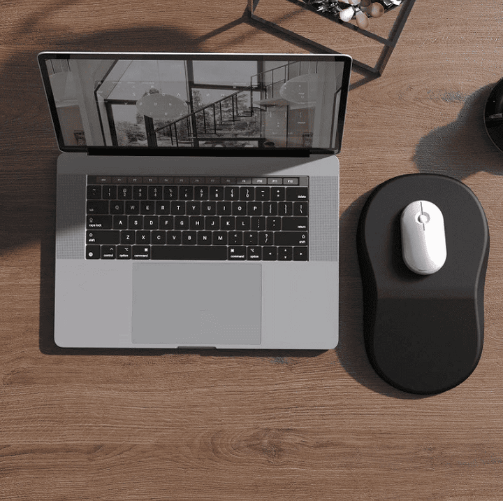 Photo prise en plongée d’un bureau en bois clair, montrant un ordinateur portable gris ouvert à gauche et, à droite, un tapis de souris ergonomique noir sur lequel repose une souris blanche.