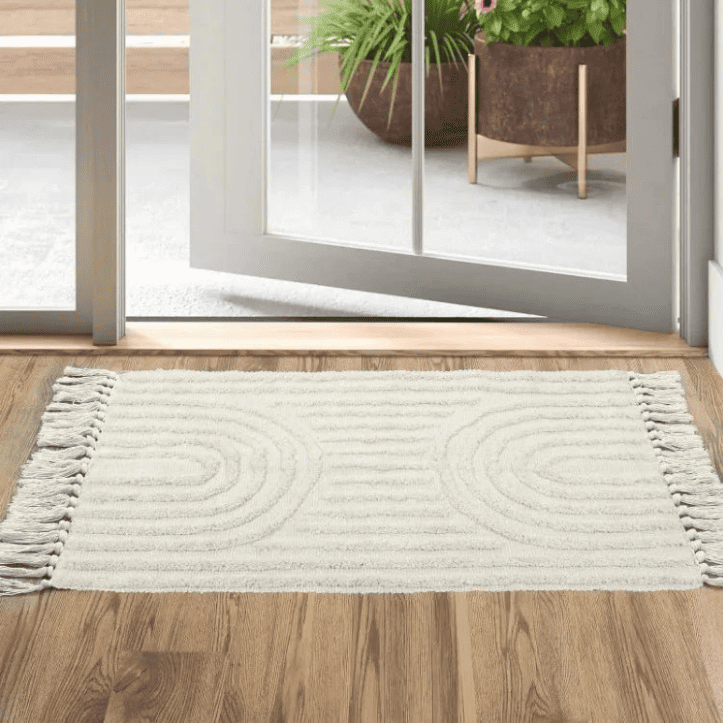 Tapis d’entrée rectangulaire crème en style scandinave avec motifs en relief et franges de chaque côté, posé devant une porte-fenêtre ouverte sur une terrasse.