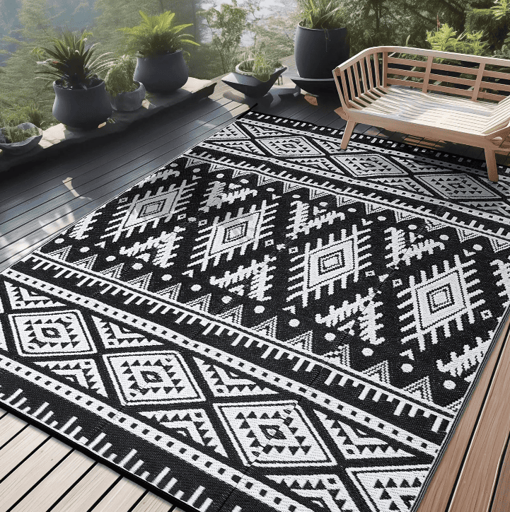 Tapis extérieur plastique motif ethnique noir et blanc posé sur une terrasse en bois avec banc et plantes