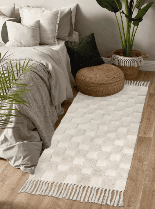 Tapis de chevet rectangulaire crème à motifs damiers en relief et franges, posé à côté d’un lit aux draps clairs, avec un pouf en jute et des plantes décoratives.