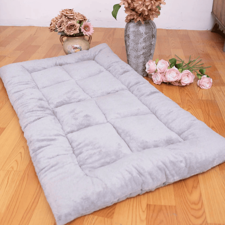 Matelas pour chien gris moelleux posé sur un sol en parquet, orné de vases de fleurs séchées et fraîches à ses côtés.