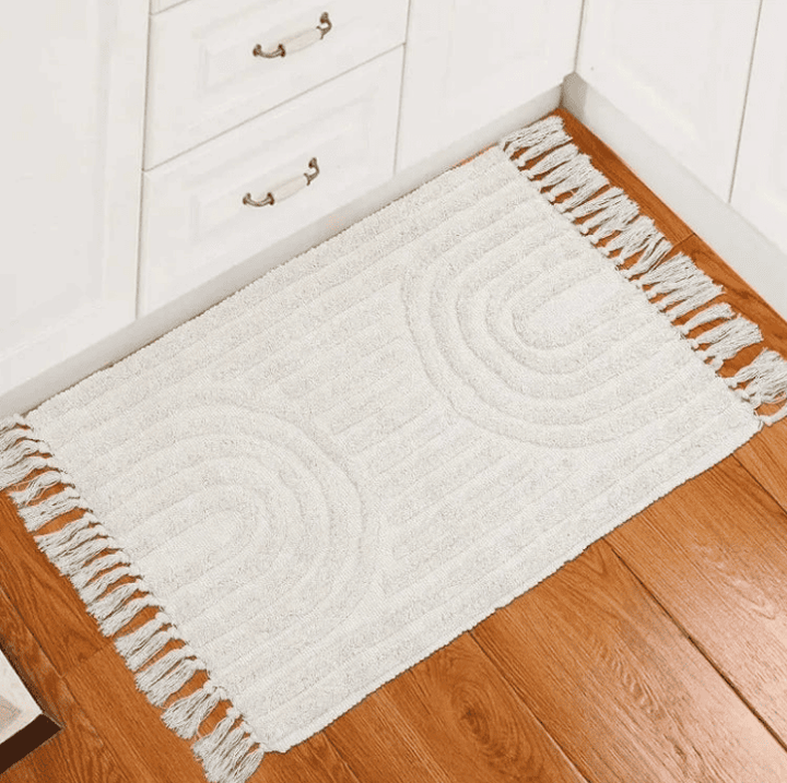 Tapis rectangulaire crème avec franges sur les côtés et motifs demi-cercles en relief, posé devant des placards de cuisine blancs sur un sol en parquet bois.