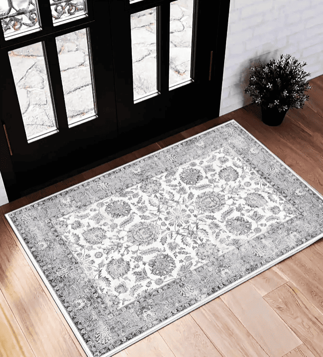 Tapis de salon lavable en machine à motif floral gris clair et blanc cassé, bordure gris moyen, posé sur un parquet bois devant une porte-fenêtre noire.