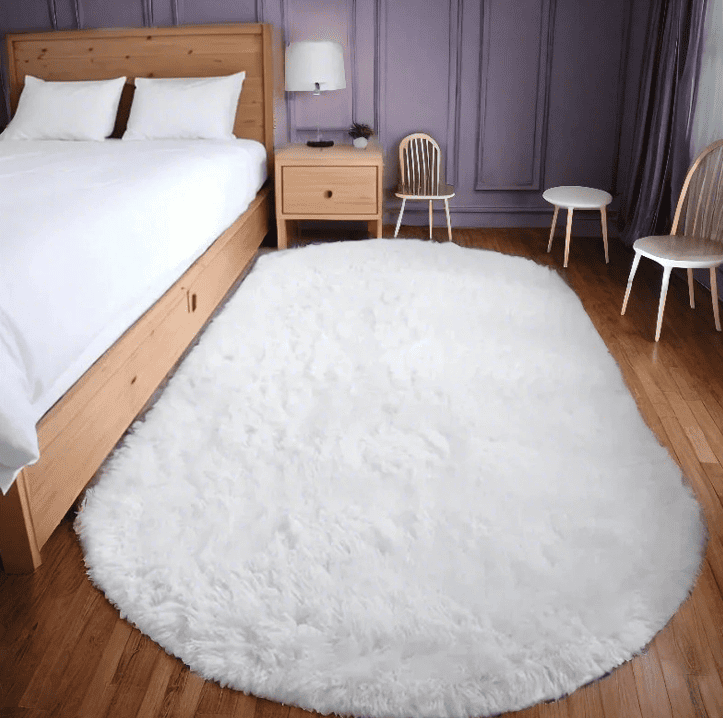 Tapis salon ovale blanc en fausse fourrure épaisse, posé sur un parquet bois devant un lit et une table de chevet