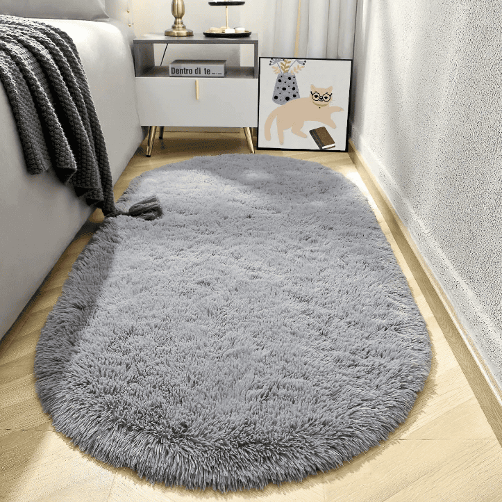 Tapis salon ovale gris clair en fausse fourrure épaisse, posé devant une table de chevet blanche et un lit, apportant une ambiance cocooning à la chambre.