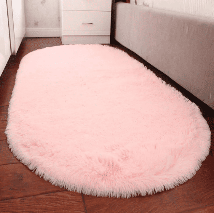 Tapis salon ovale rose pastel en fausse fourrure, posé le long d’un lit dans une chambre, créant une touche cosy sur un sol en parquet foncé.