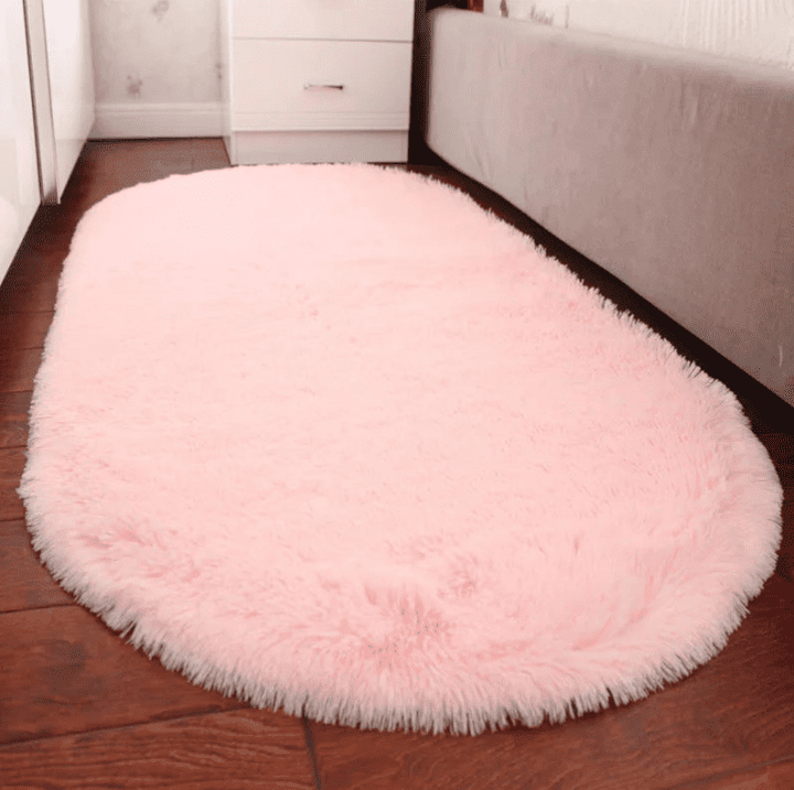 Tapis salon ovale rose pastel en fausse fourrure, posé le long d’un lit dans une chambre, créant une touche cosy sur un sol en parquet foncé.