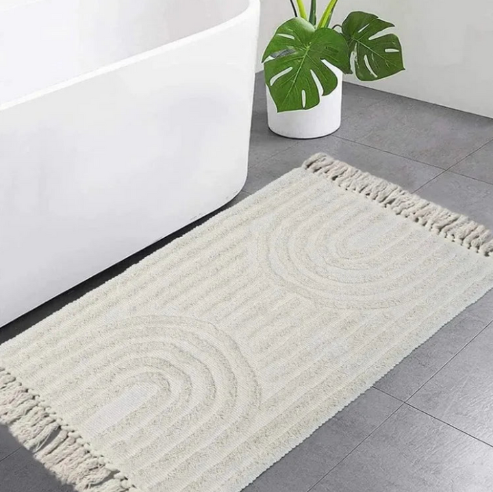 Tapis rectangulaire scandinave couleur crème avec franges aux extrémités et motifs demi-cercles en relief, posé au sol devant une baignoire blanche sur un carrelage gris.