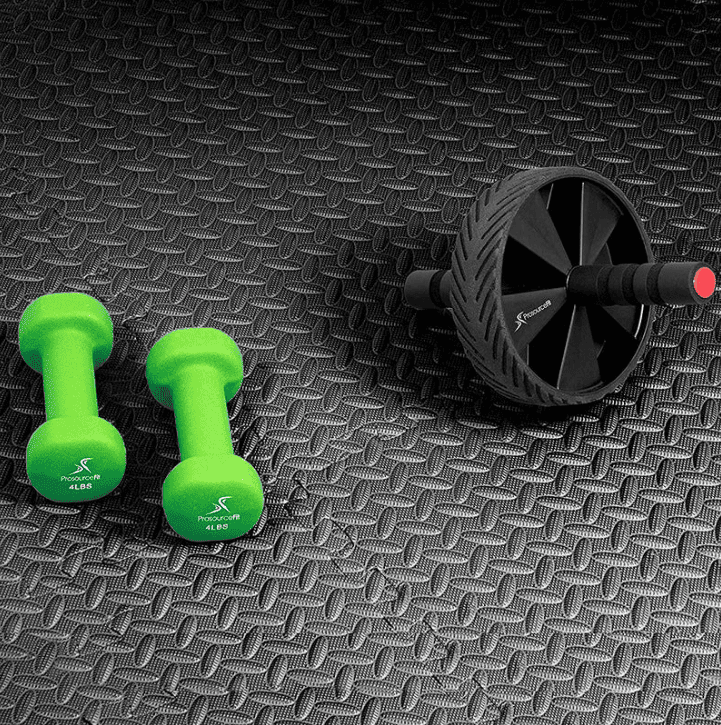 Tapis de sport en salle noir texturé, avec haltères verts et roue abdominale posés dessus pour démontrer l’adhérence et la protection du sol.